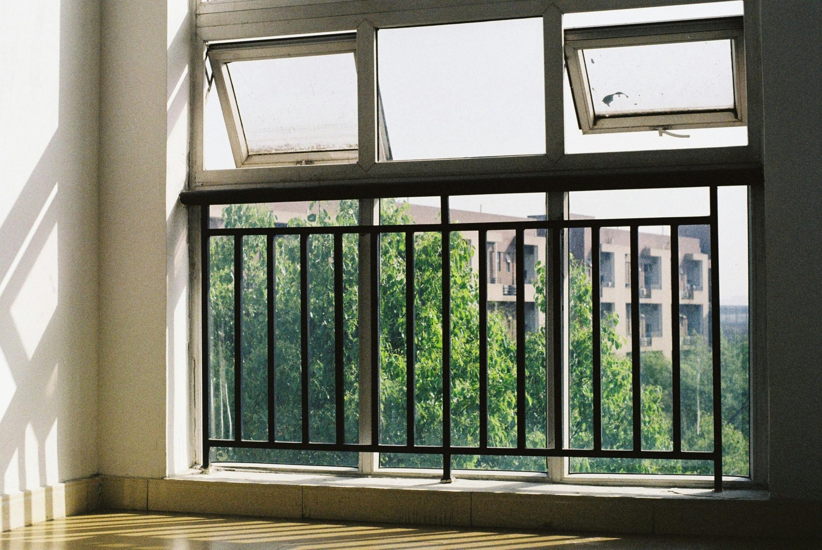 A sunlit room with open windows and metal bars, overlooking greenery.