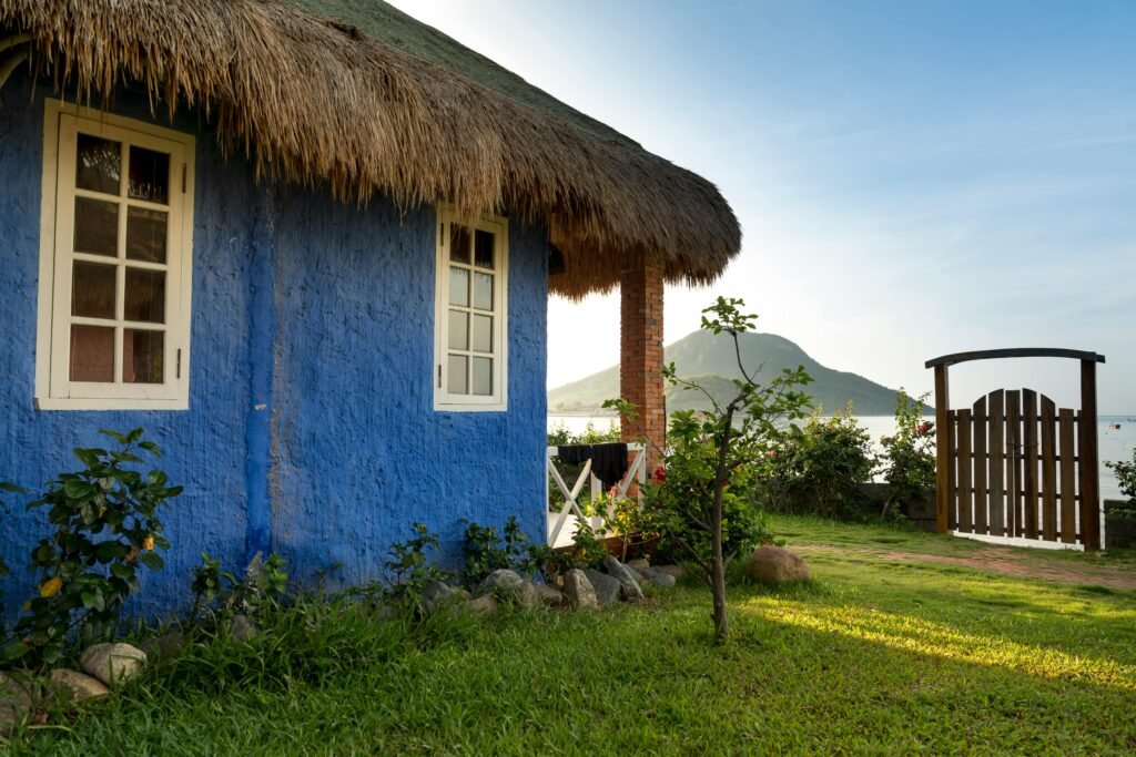 Rustic blue cottage with thatched roof by a serene lakeside and mountain view.